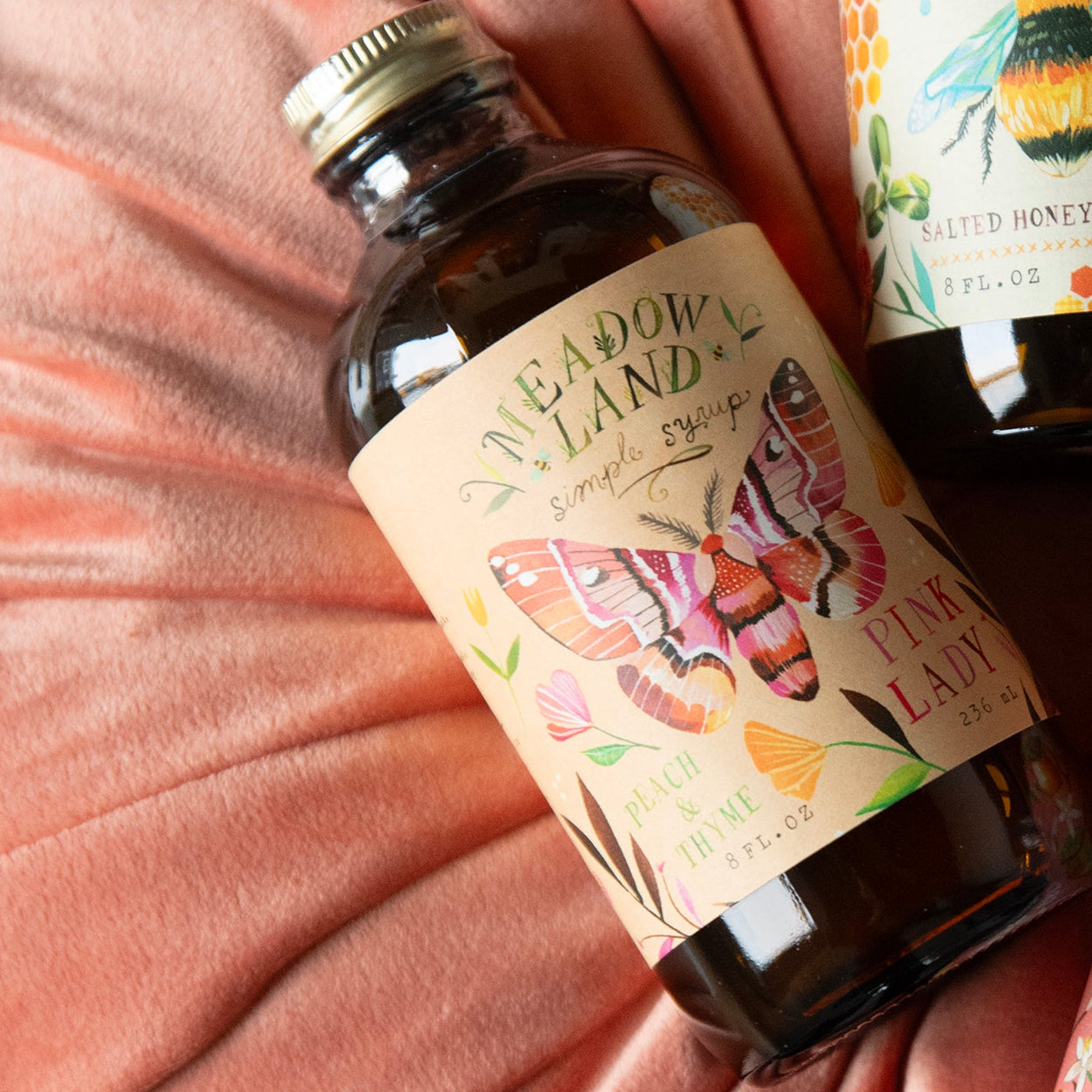 Meadowland simple syrup bottle with illustrated label, arranged on a gathered dusty pink velvet fabric surface.